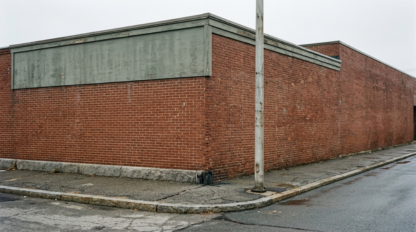 A plain street corner with a low granite kerb, a bare pole and an empty painted wall above a single story building, in flat gray daylight