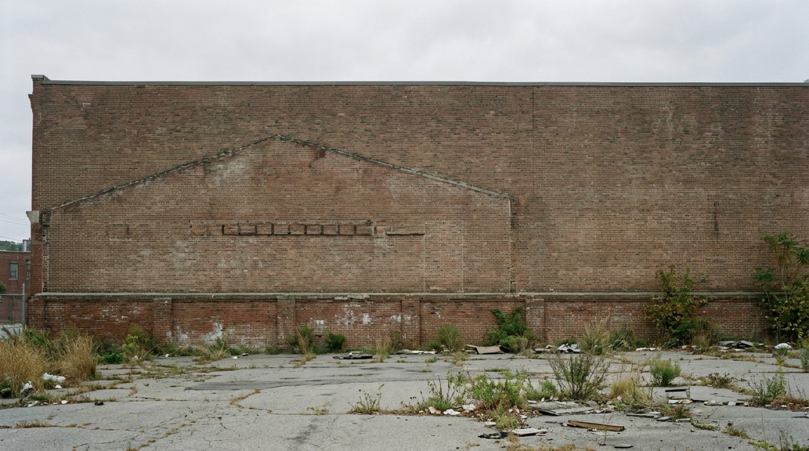 An empty lot of cracked asphalt and grass with a low brick foundation wall still standing along one edge, and the blank party wall of the next building rising behind it